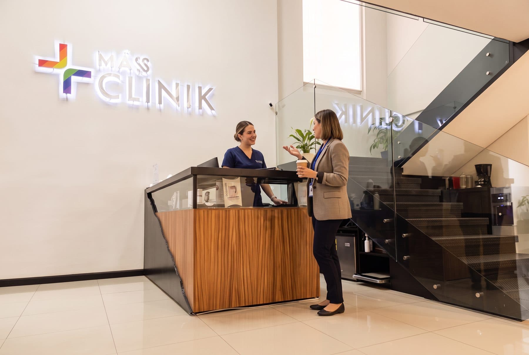 Recepción de +Máss Clinik con el logo iluminado: una recepcionista atiende a una profesional que llega con un café en la mano